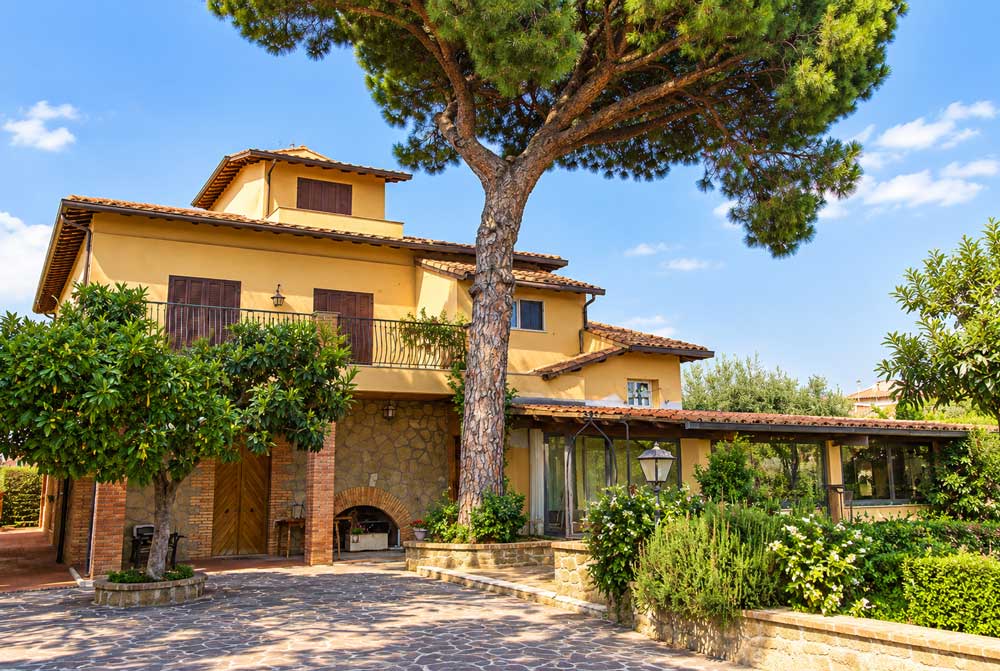 Facciata del casale con pino domestico, balcone e cortile lastricato